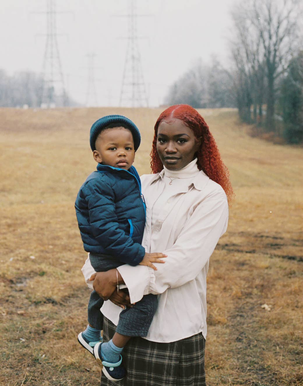 A woman holding her son close in a field of dry grass with electrical towers in the background