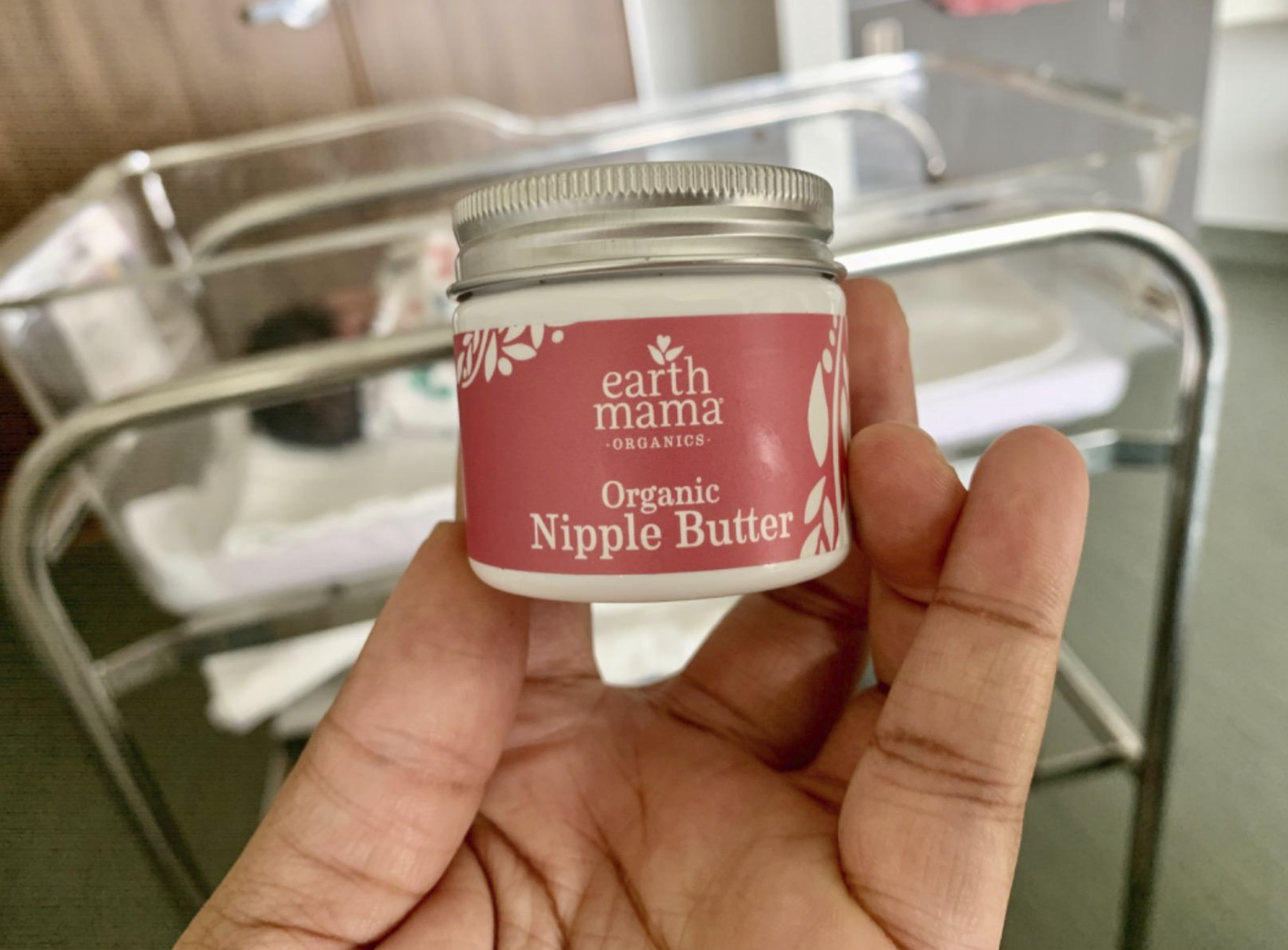 reviewer holding a jar of the nipple butter in front of their baby&#x27;s nursery crib in the hospital