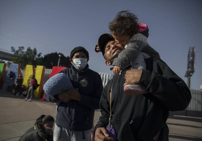 A man smiles with a young child on his shoulder as another adult stands in the background