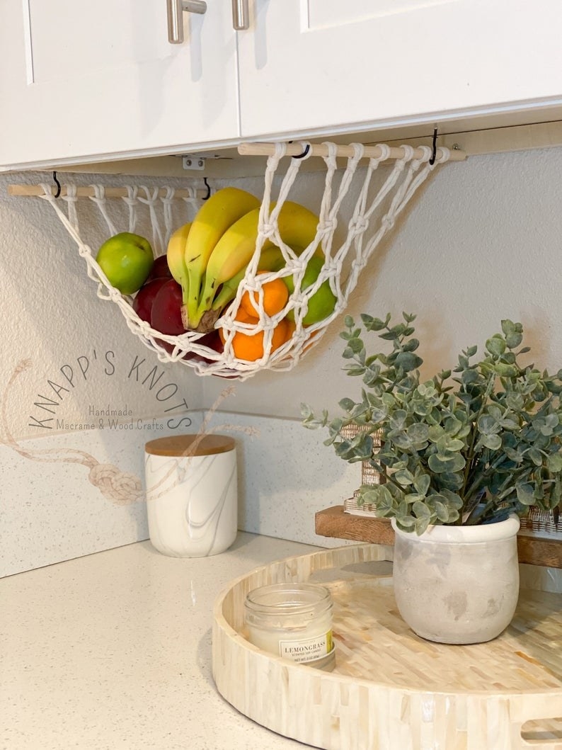 the fruit hammock with white rope hanging underneath a kitchen counter with bananas, apples, and oranges inside 