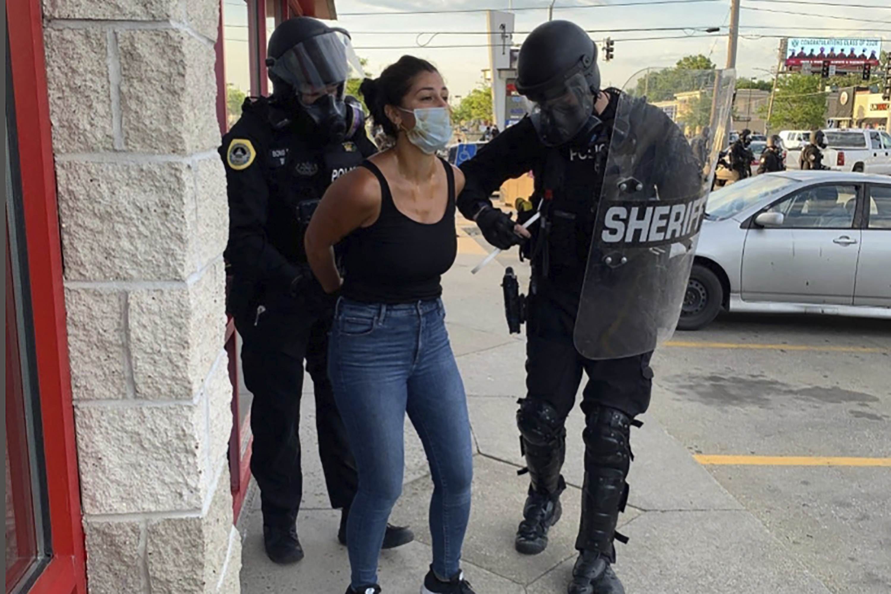 A woman wears a face mask and has her hands behind her back as she is arrested by police officers in armor and gas masks