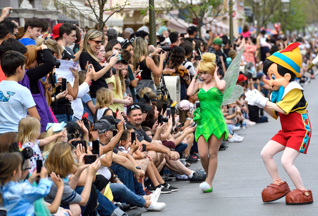 A large crowd lines a Disneyland street to watch the parade and interacts with Tinker Bell and Pinnochio