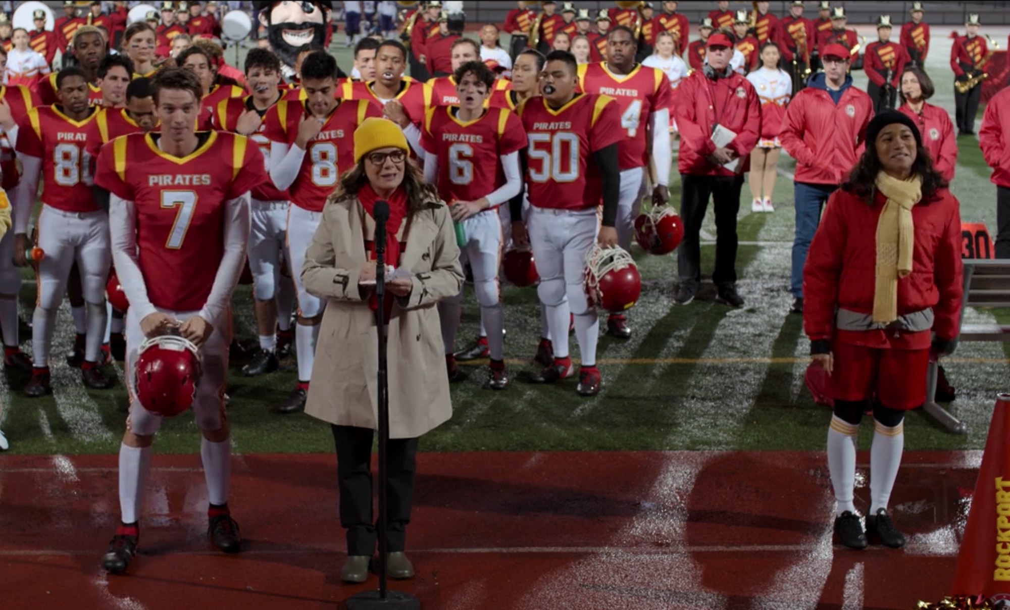 High school&#x27;s football team stands on the field behind the principal at a microphone