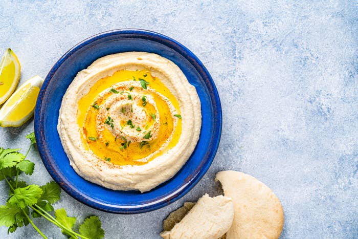 A bowl of hummus, topped with a light coriander garnish; lemon, bread and coriander sit around the bowl 