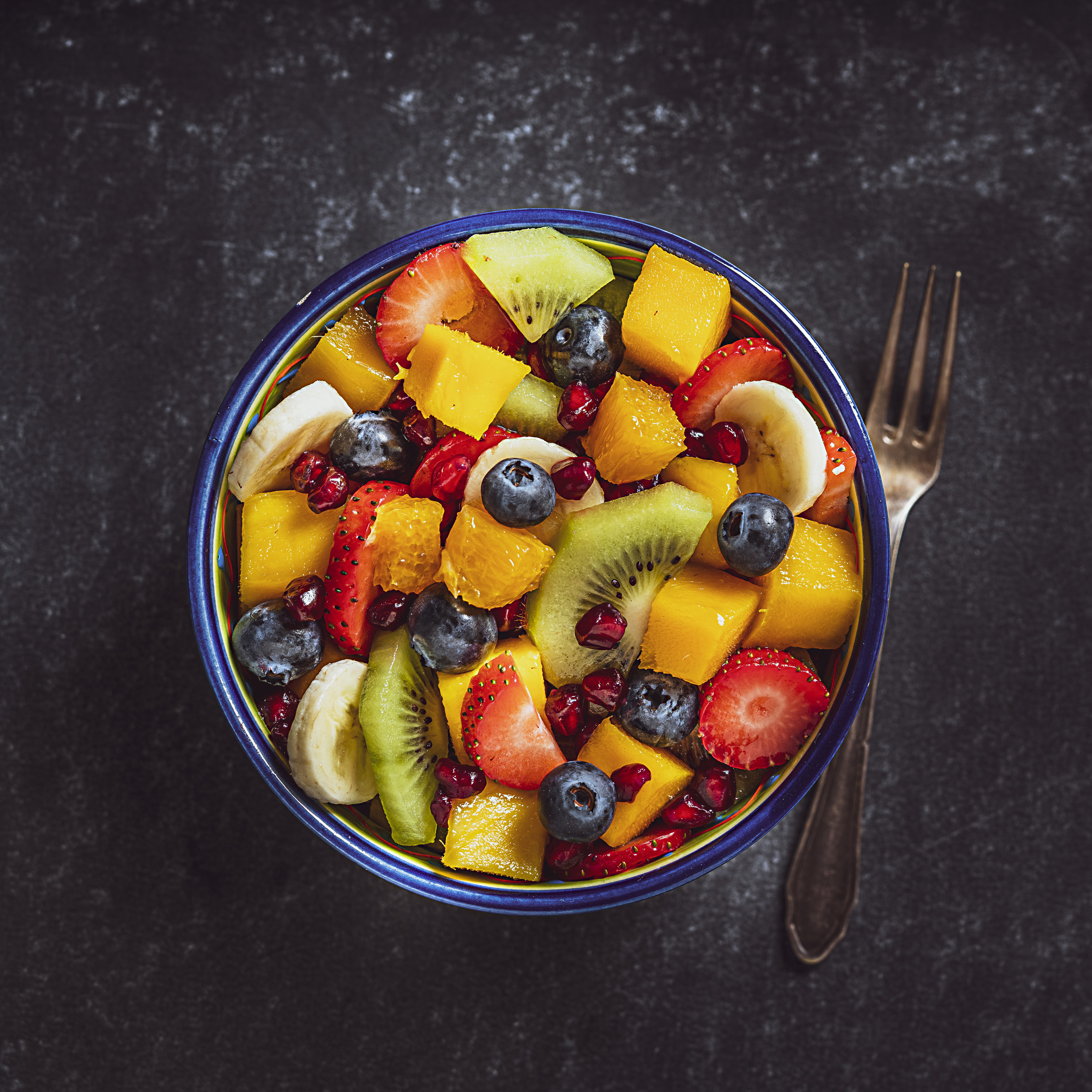 Homemade salad bowl shot from above on dark table with a fork beside the bowl; fruit included are mango, orange, kiwi, strawberry, banana, pomegranate and blueberries