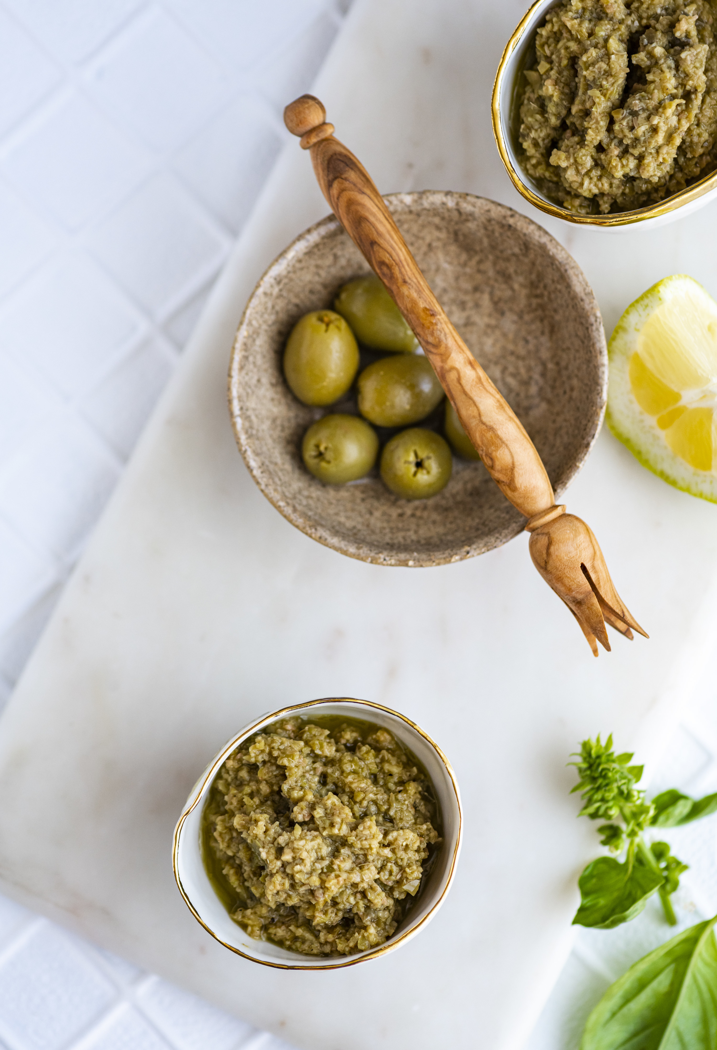 A bowl featuring a handful of olives and two bowls featuring olive tapenade all placed on a chopping board with a lemon in the background