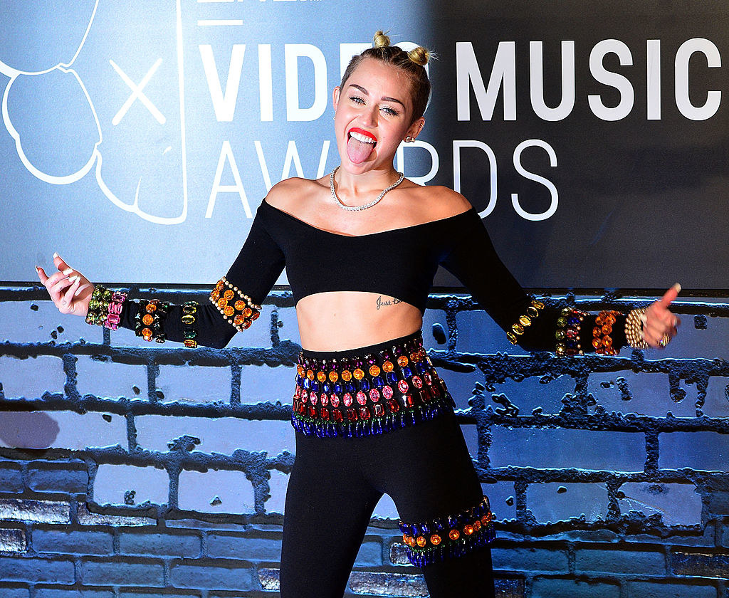 Miley posing on a red carpet and sticking her tongue out while wearing a long-sleeved crop top and matching bejeweled pants