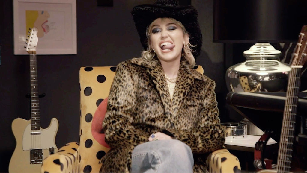 Miley sitting in a chair, wearing a leopard print coat and jeans, and sticking her tongue out