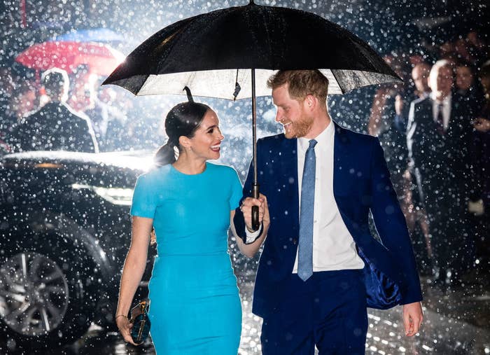 Harry and Meghan smiling at each other as they walk through the rain and hold an umbrella together