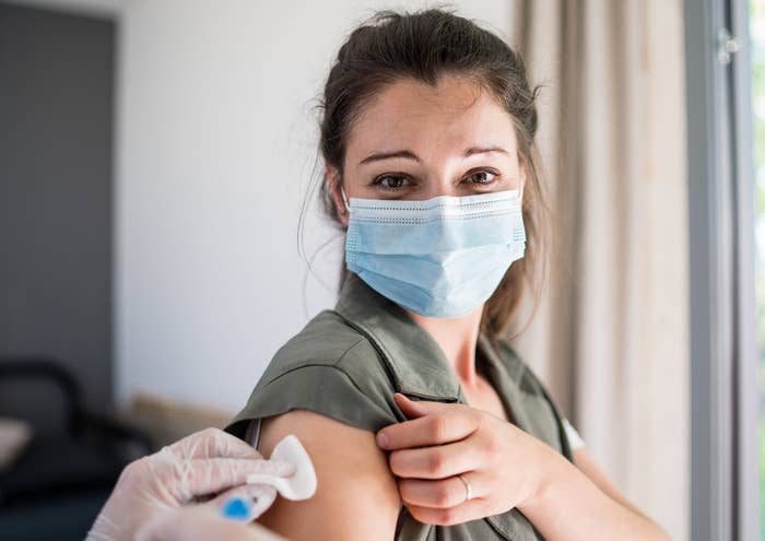 A woman wearing a disposable face mask pulls up her sleeve as a gloved hand wipes her arm with a cloth