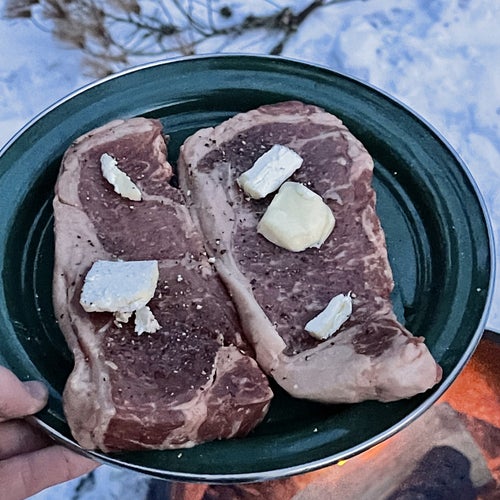 Two uncooked steaks covered in seasoning and butter above a campfire 