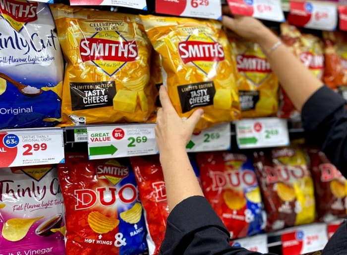 Hands reaching for a packet of &quot;Tasty Cheese&quot; Smith&#x27;s chips on the shelf amongst other Smith&#x27;s flavours