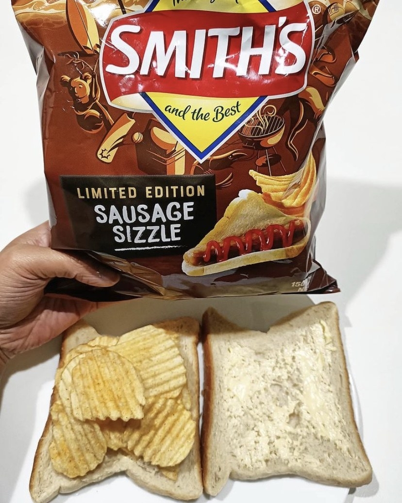 Two pieces of buttered white bread with the left hand side piled high with chips; a hand holding a packet of &quot;Sausage Sizzle&quot; chips hover above