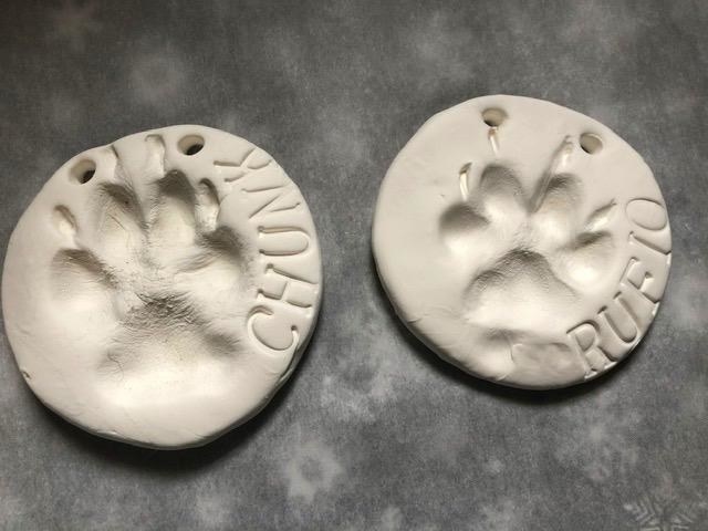 Two ornaments, made from white clay, with each dog&#x27;s name stenciled on the ornament beside their paw print