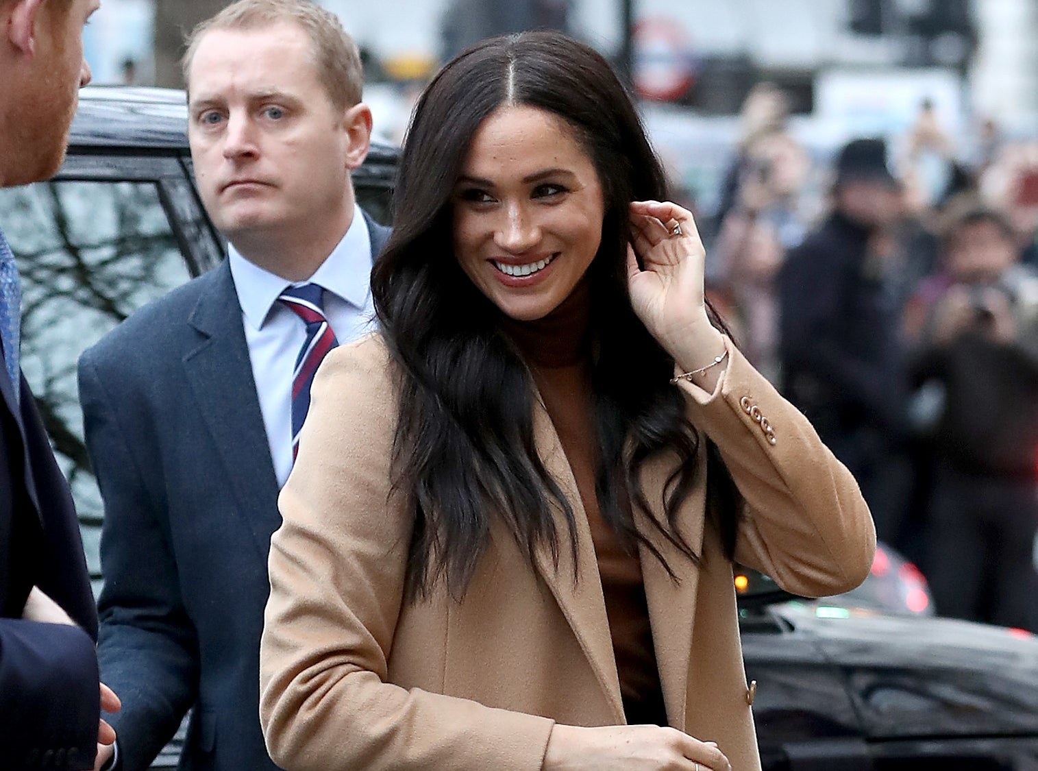 Meghan smiles in a beige wool coat on the way to an event while a crowd stands behind her