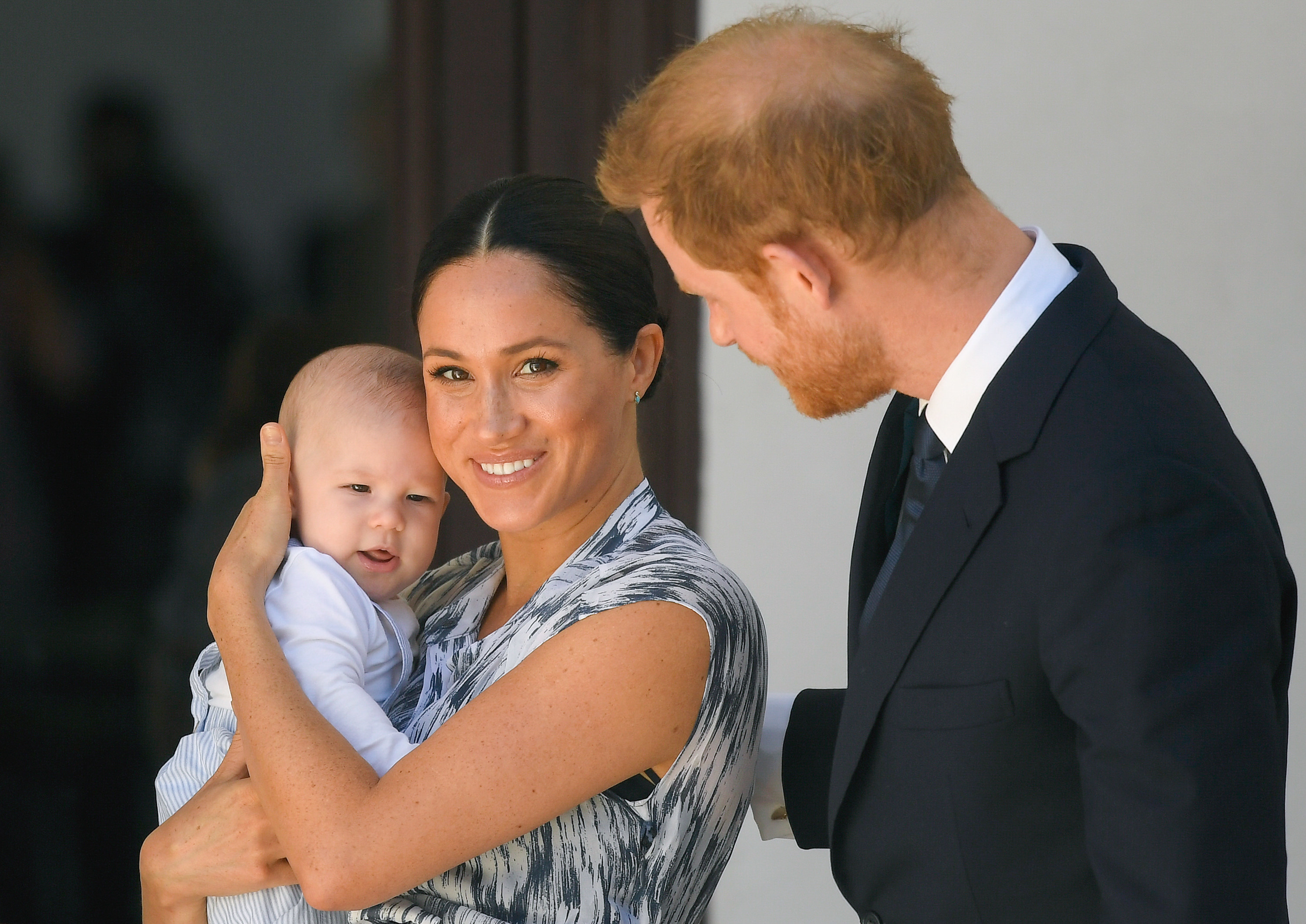 Meghan smiles while holding Archie and Harry puts his arm around her