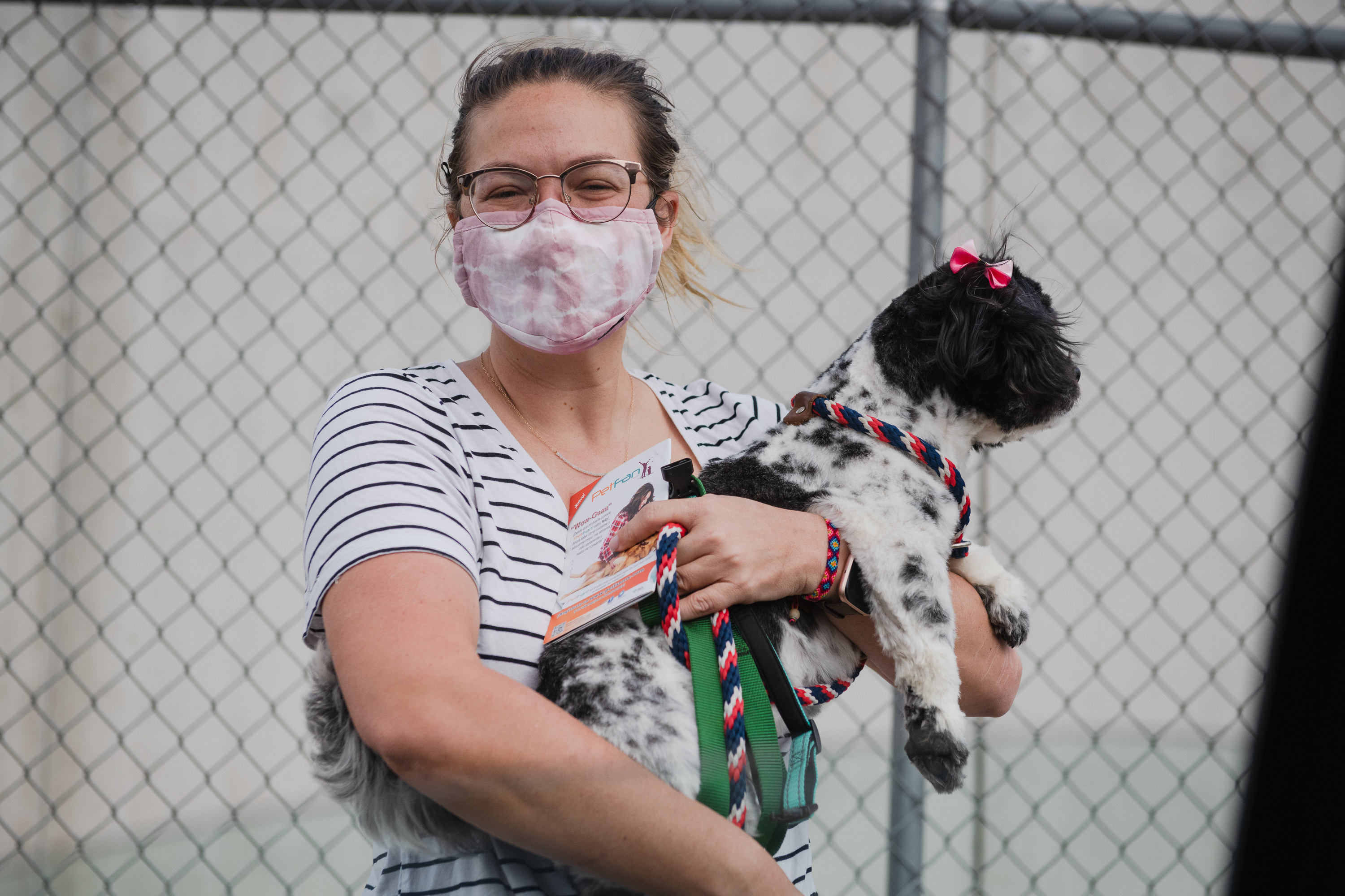 A woman wearing a face masks smiles widely and holds a small dog with a bow above its ear