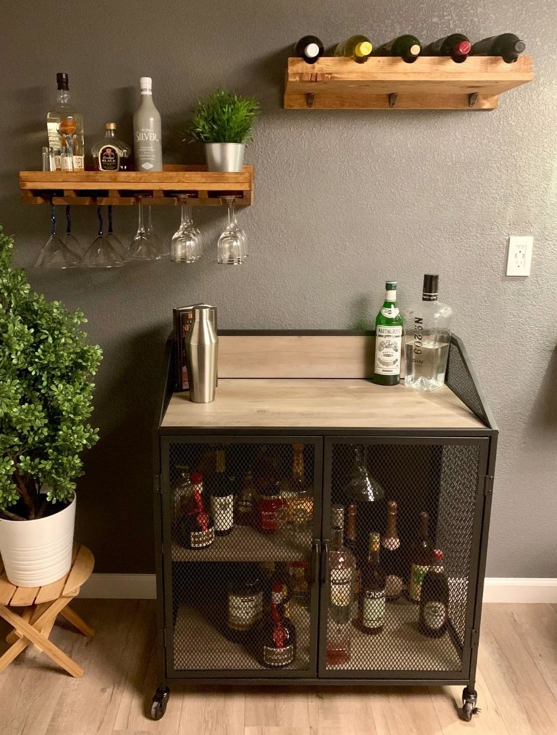 reviewer image of the Walker Edison Malcomb Urban Industrial Metal Mesh Double Door Rolling Bar Cabinet with several bottles of alochol in the cabinet space and a bottle of gin on its top surface