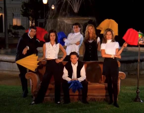 The Friends cast sitting on an orange couch in front of a fountain