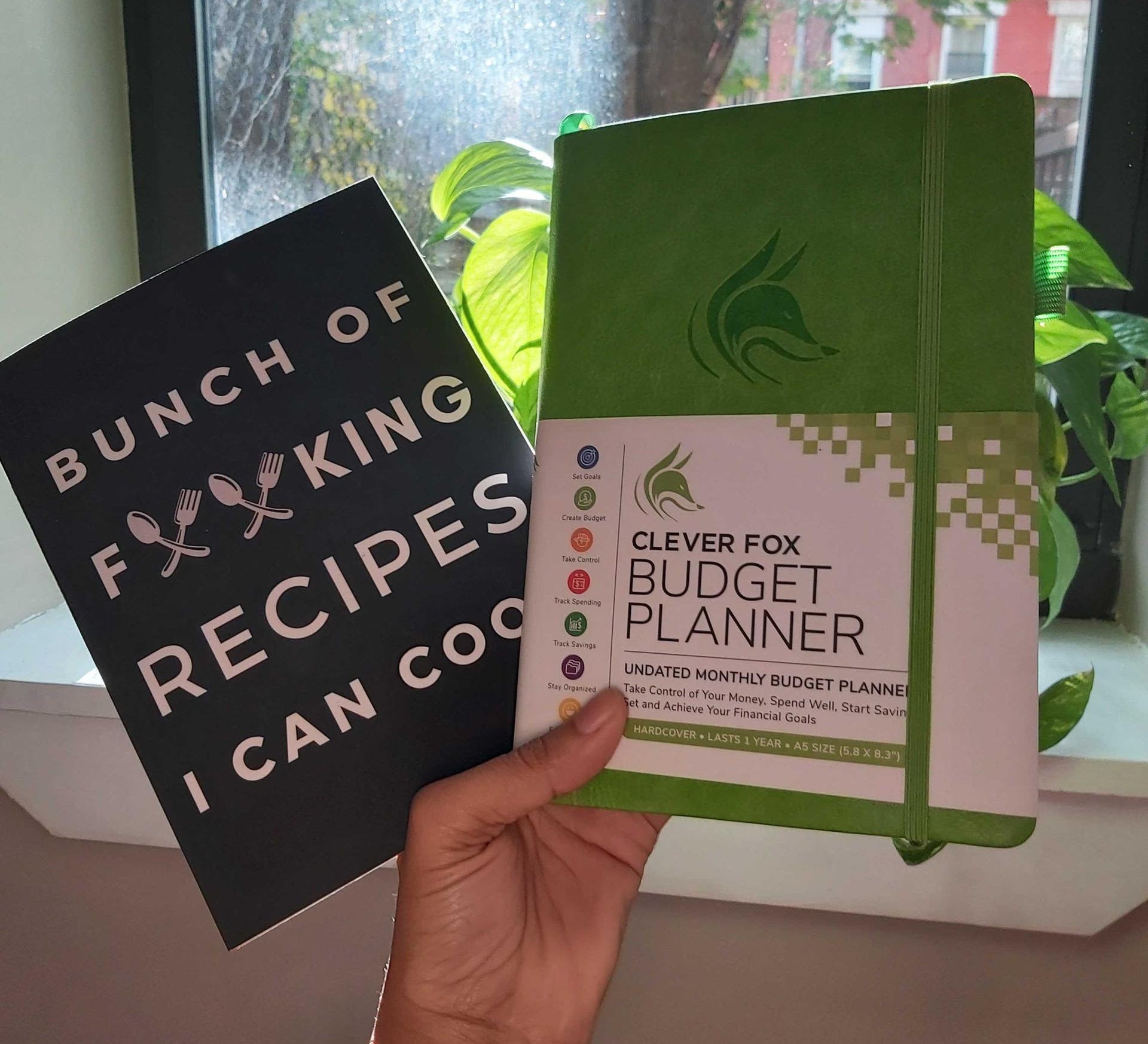 buzzfeeder holding the budget planner and a recipe book that reads &quot;bunch of forking recipes I can cook&quot; with forks in the middle of the word &quot;forking&quot;