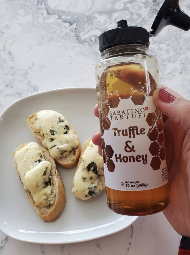 reviewer's hand holding bottle of truffle handy in front of bruschetta with cheese on it