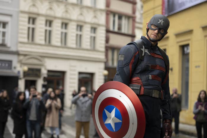 John Walker looking angry, standing in a European town square, holding Captain American's shield