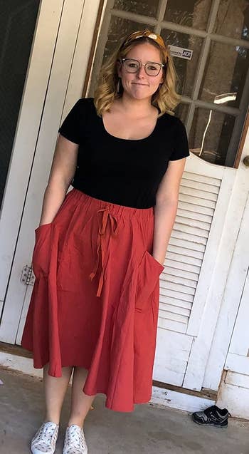 a reviewer wearing sneakers and a black T-shirt tucked into the red midi skirt with their hands in the two side patch pockets 
