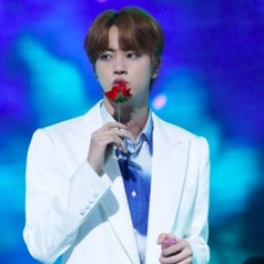 Jin has light brown hair and wears a white suit, he holds a fake rose and is kissing it; his other hand holds a pink sparkly mic