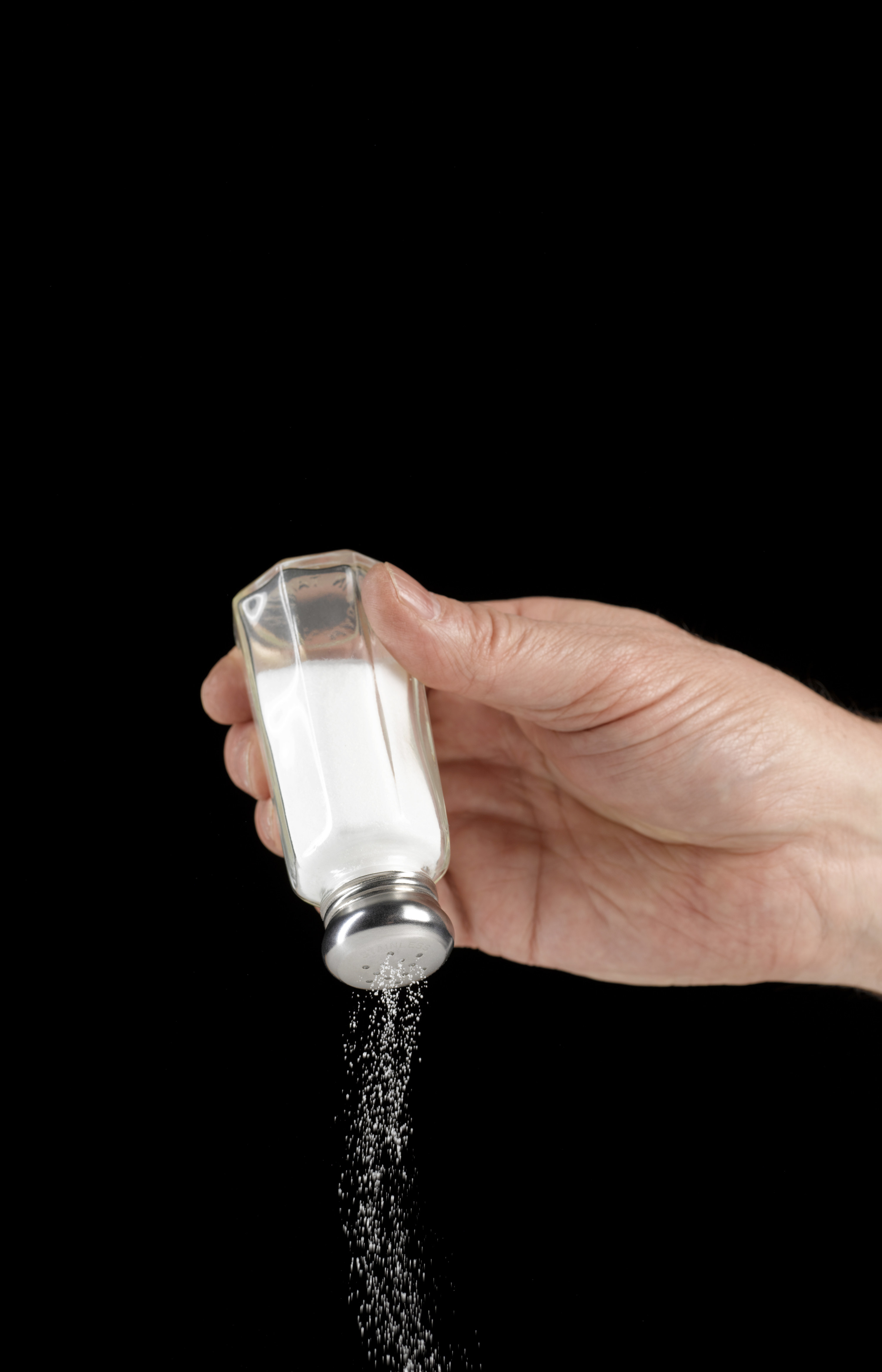Hand holding a salt shaker upside, forcing salt to be shaken out. Photo is set against a dark black background.