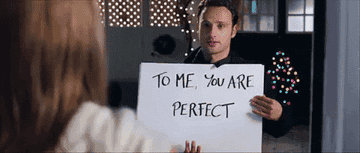 Andrew as Mark holding up large cards that say, &quot;To me you are perfect and my wasted heart will love you&quot;