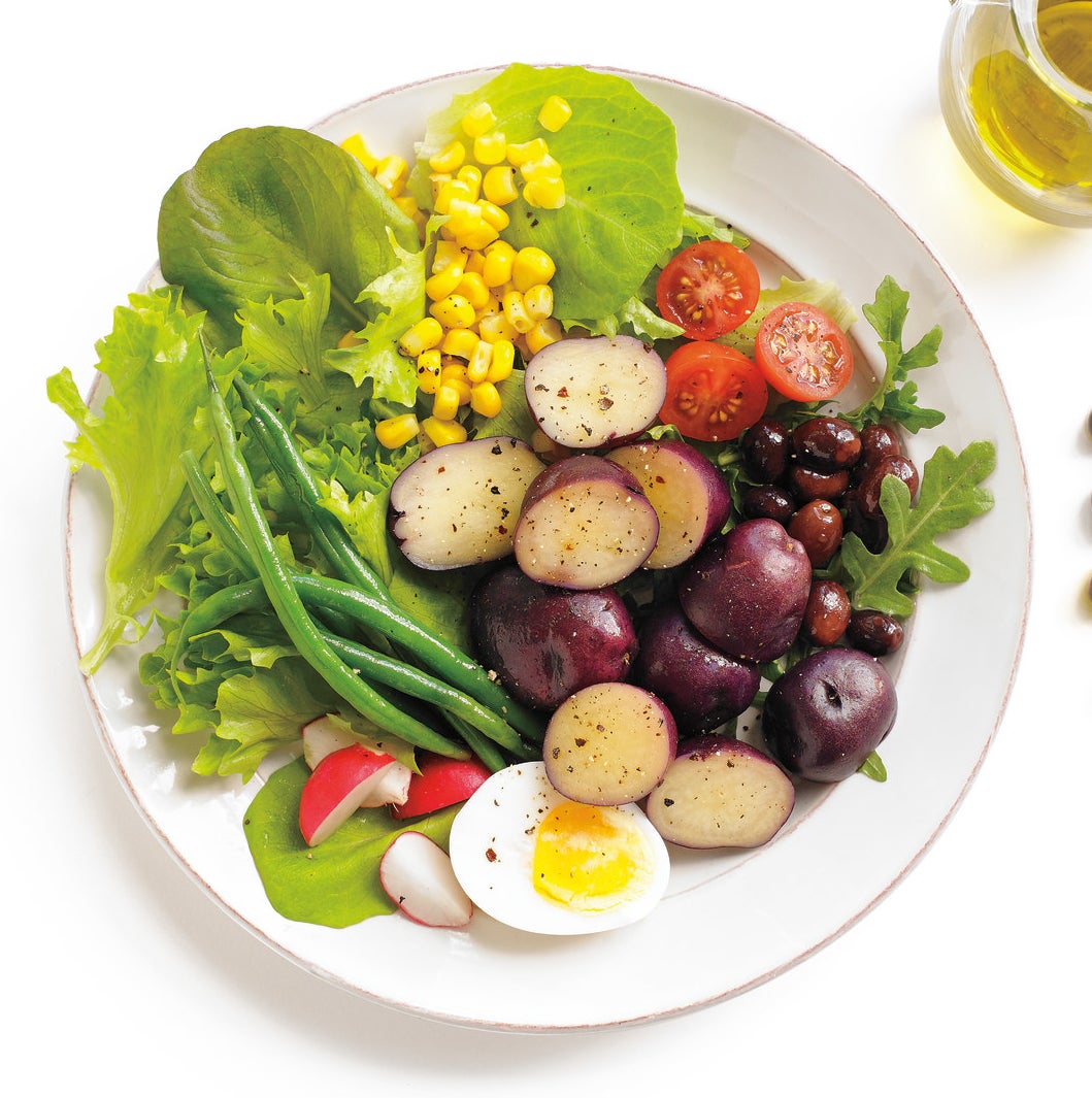 An arranged niçoise salad: little potatoes, half a boiled egg, olives, cherry tomatoes, green beans, sweet corn, and radishes on a bed of lettuce.