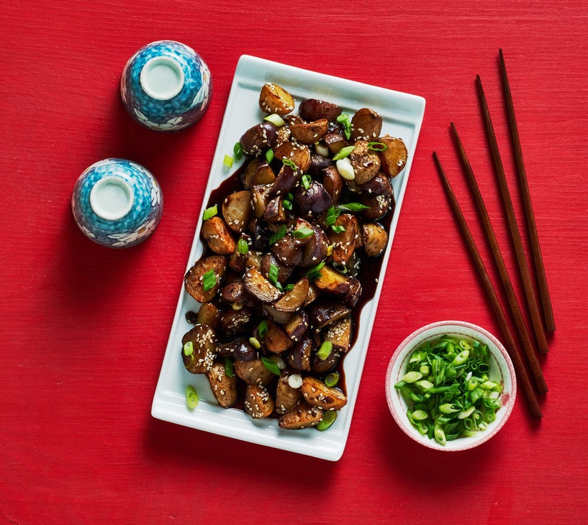 A serving plate of sesame little potatoes with a side of green onions. Two pairs of chopsticks are on the table.