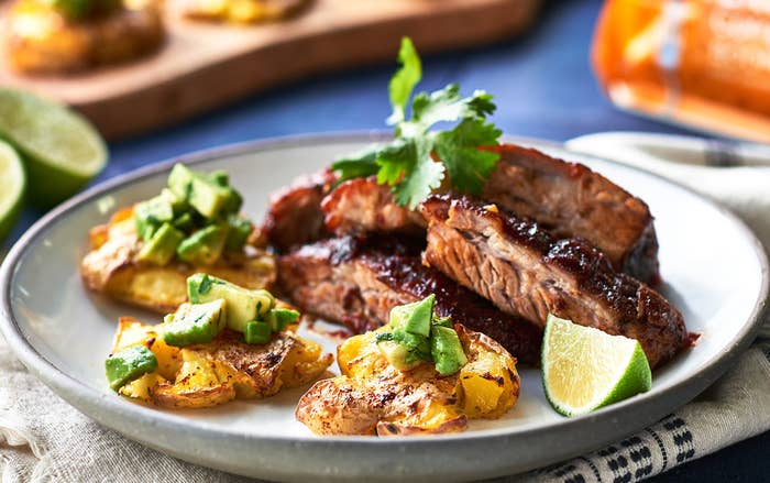 A plate of BBQ served with smashed little potatoes, garnished with cilantro, diced avocado, and a wedge of lime.