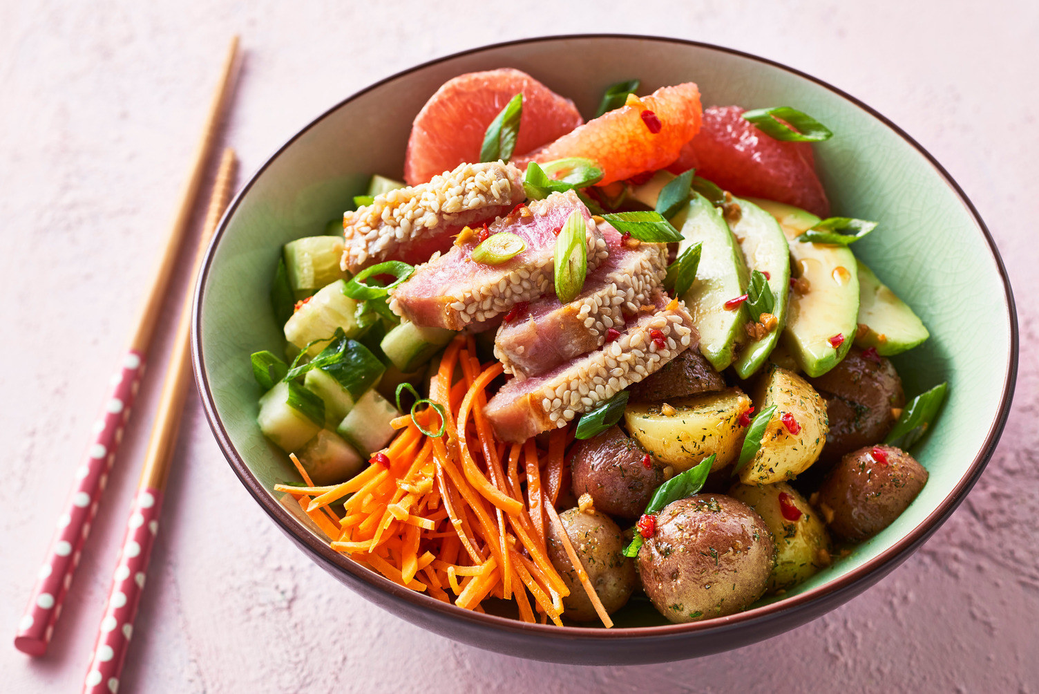 A bowl of poke with sesame-crusted tuna, julienned carrot, avocado, chopped cucumber, grapefruit, roasted little potatoes, and a spicy dressing. 