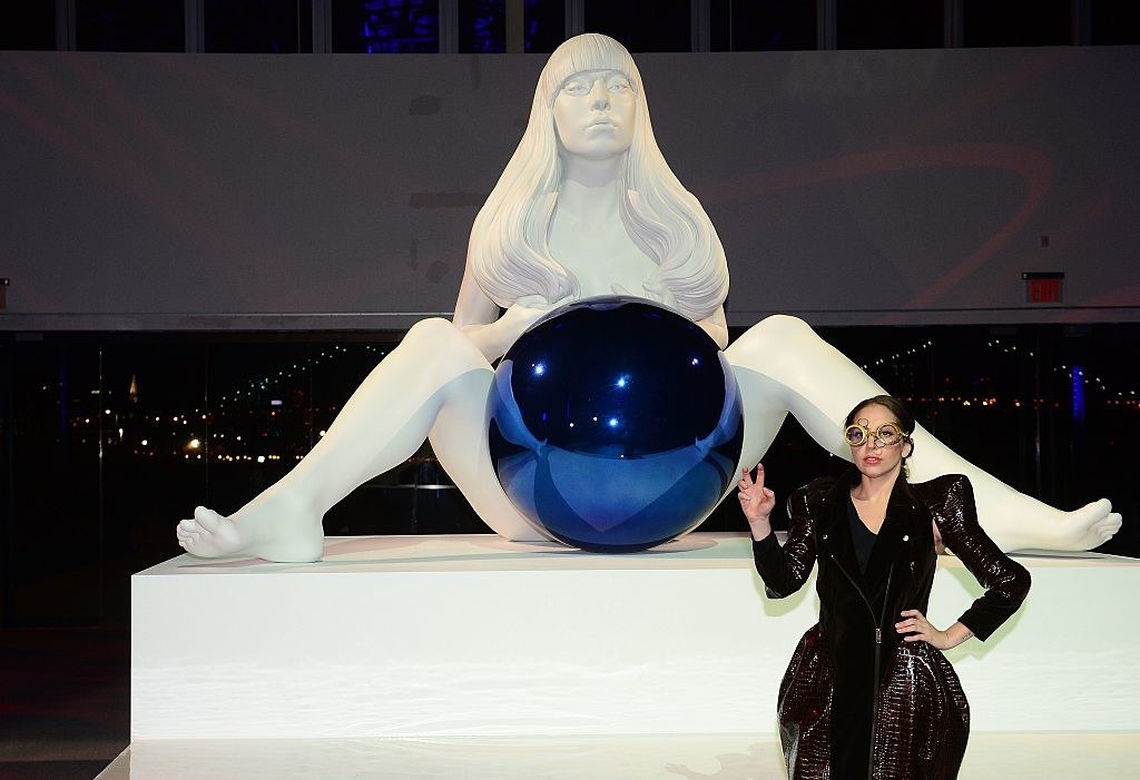 Gaga standing in front of a sculpture of herself in which her legs are spread and a giant blue ball sits in front of her