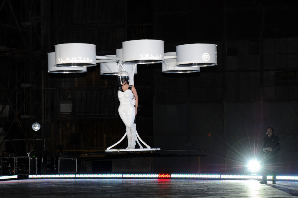 Lady Gaga being carried by six propellers that are attatched to a white dress shaped like a body