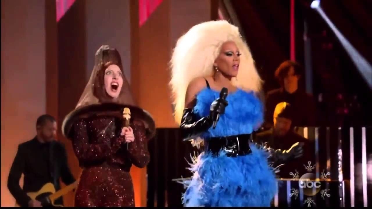 Gaga singing wearing a brown headpiece that resembles a condom while next to RuPaul