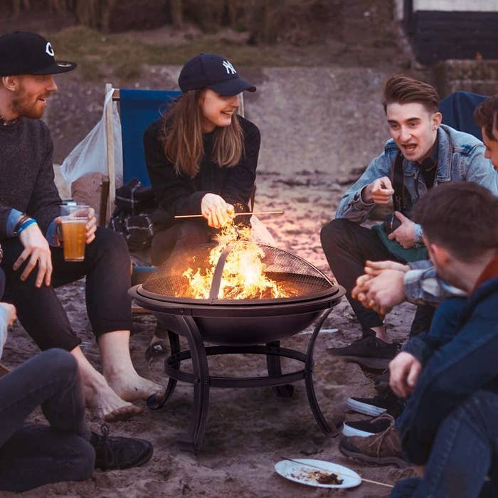 five smiling models sit around fire pit, which has a big fire, on the beach