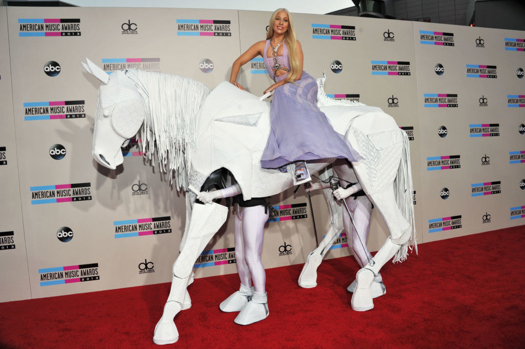 Gaga riding on a white mechanical horse operated by two men in latex