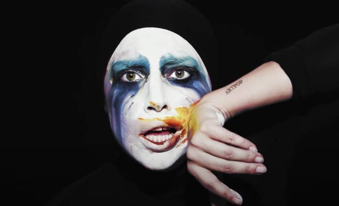 Lady Gaga in white face makeup and colors running down her face as she wipes her mouth and shows off the Artpop tattoo on her forearm