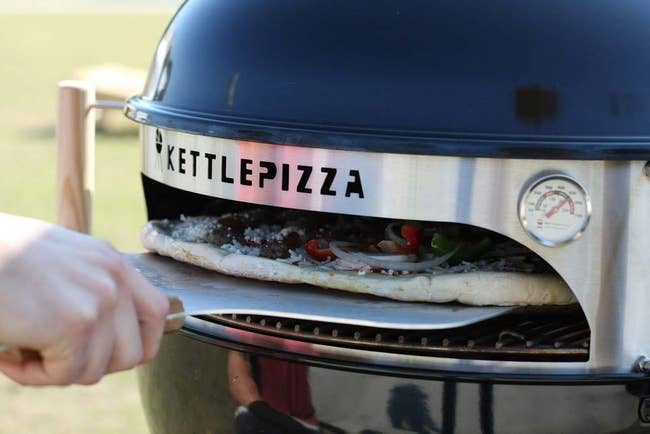 A hand inserting a pizza into a grill that's been converted into a pizza oven