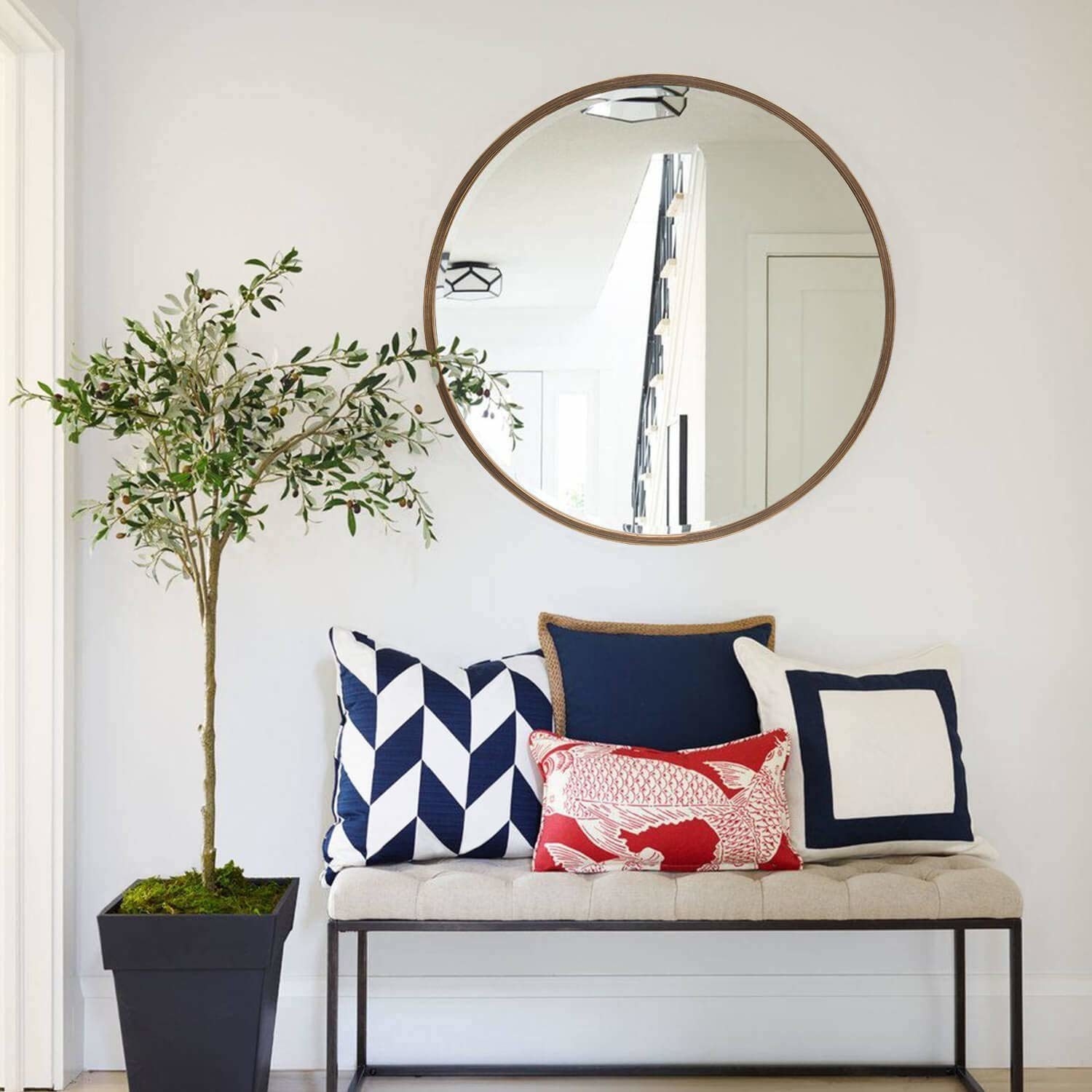 A circular mirror with a brown border hung up on a wall. There&#x27;s a white sofa with some colourful cushions below the mirror, and there&#x27;s a tall plant next to it.