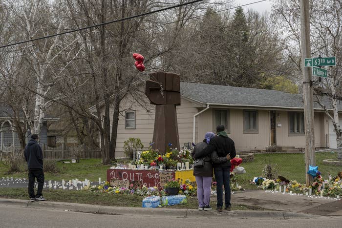 Two people stand side by side by a large clenched-fist monument surrounded by flowers and candles at a street corner