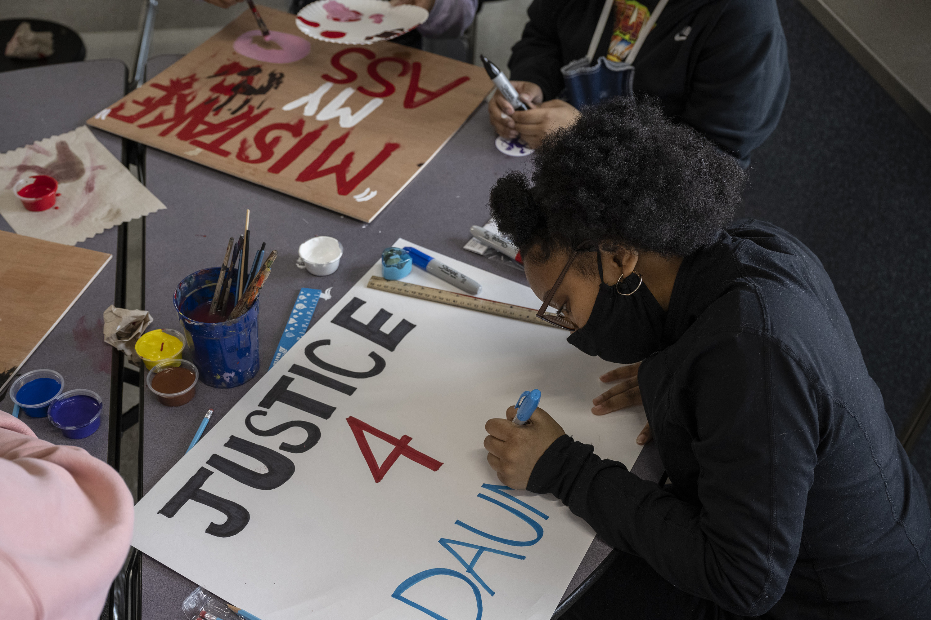 A Black student wearing a face mask sits at a table with art supplies and fills in a protest sign that reads &quot;Justice for Daunte&quot;