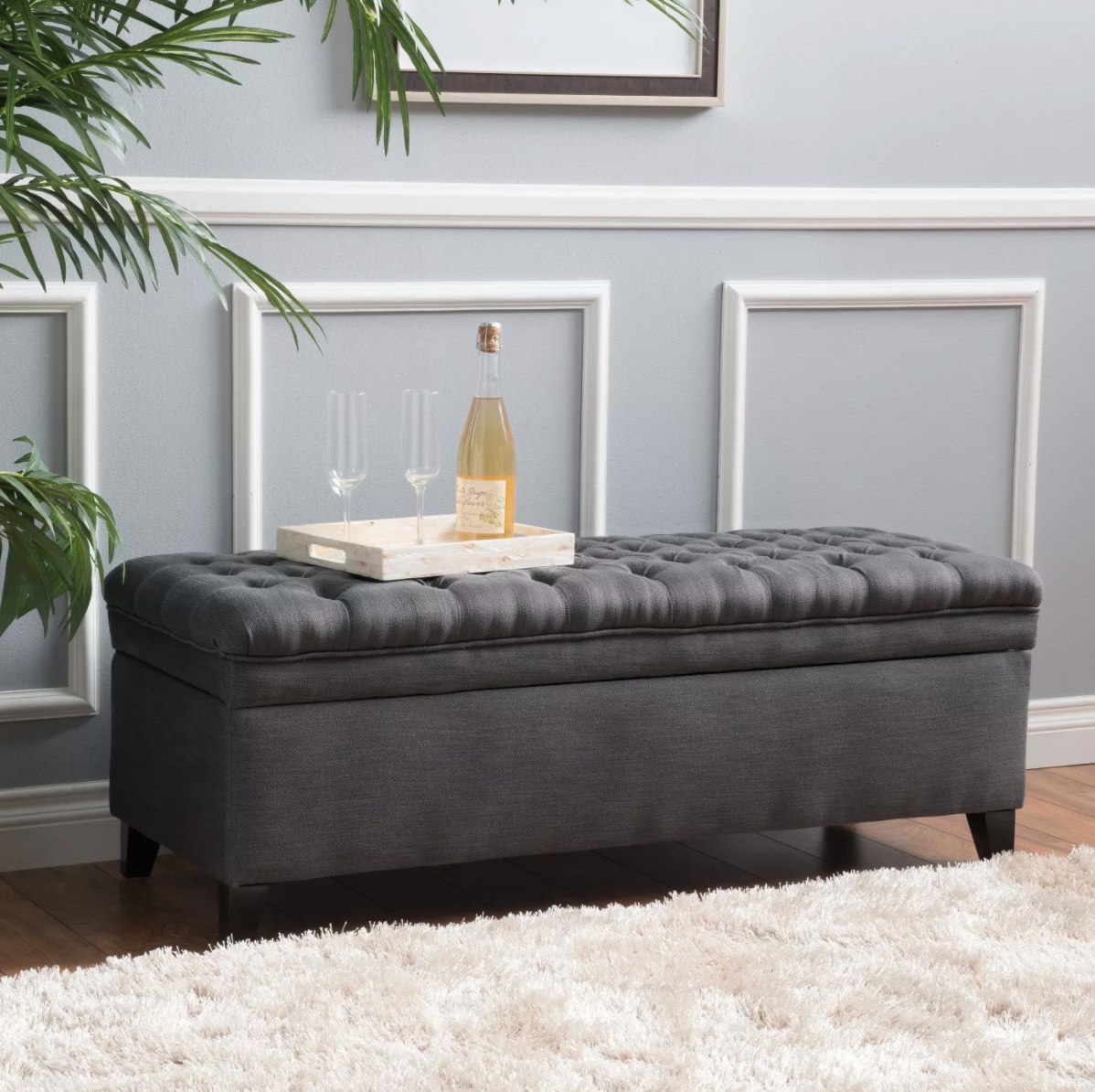 The storage ottoman in gray with a tray with flutes and champagne on top