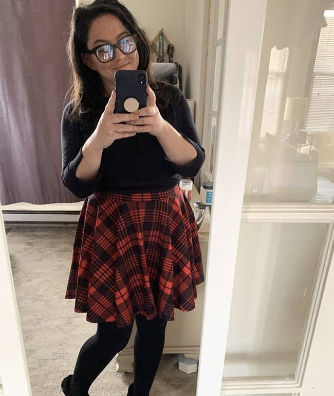 A reviewer wearing a black top, red plaid skirt, and tights