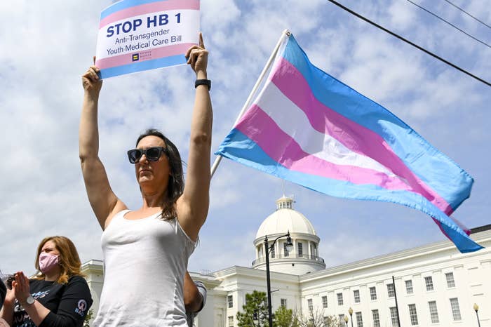 A person stands in front of a trans pride flag and holds a sign reading &quot;Stop HB 1 Anti-Transgender Youth Medical Care Bill&quot;