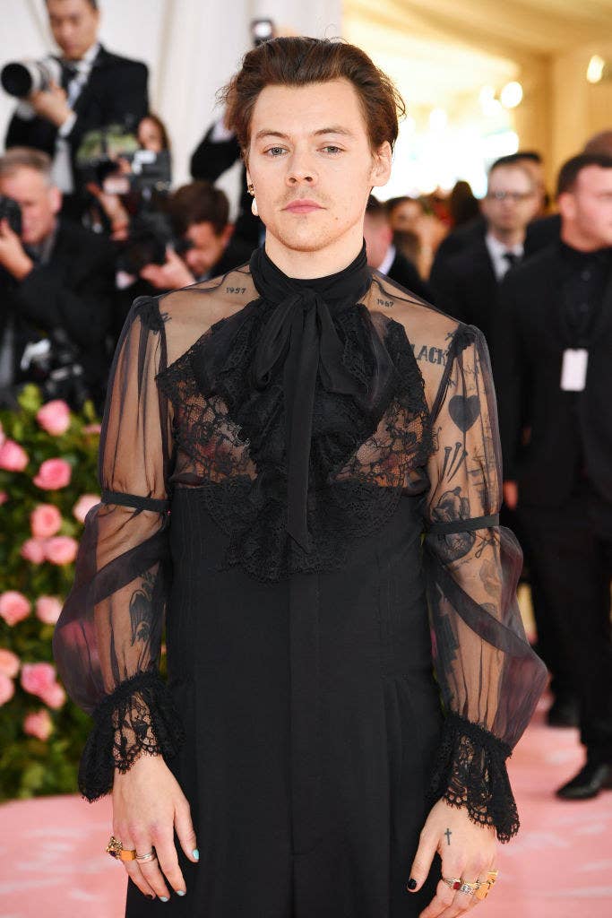 Harry at the Met Gala wearing a see-through blouse with lace, a single pearl drop earring, and several rings on his fingers