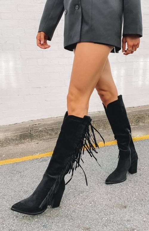 model wearing the pointed toe, just-below-the-knee height boots in black with fringe on the sides