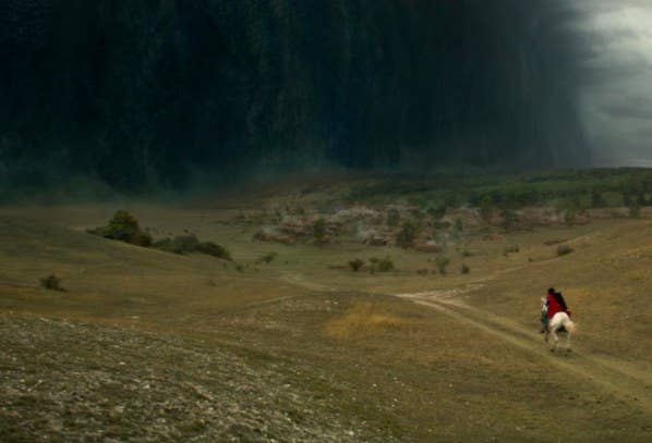 Someone on horseback rides through a green countryside; a dark shadowy wall of clouds called the Fold is ahead of them